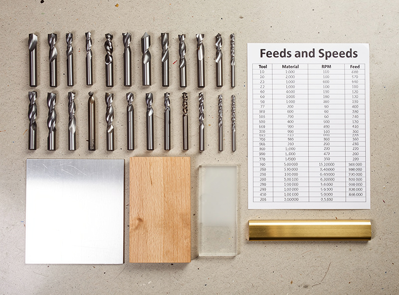 Top down view of various CNC end mills and material samples on a workbench with feeds and speeds reference chart visible
