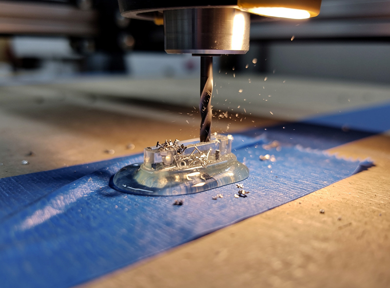 A small part bonded to a CNC spoilboard with the tape and CA glue method while an end mill cuts detail