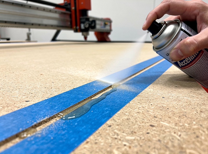 CA glue and accelerator being applied between two painter's tape layers to bond a part to a CNC spoilboard